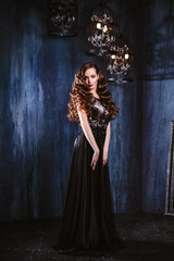 Young woman with long curly hair and makeup in evening long luxury dress, posing in a dark interior room. fashion beauty portrait