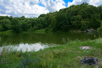 The waterfall on the river flows through and over the rocks covered with lichen and moss against a background of green vegetation and a blue sky.