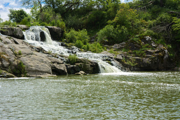 The waterfall on the river flows through and over the rocks covered with lichen and moss against a background of green vegetation and a blue sky.