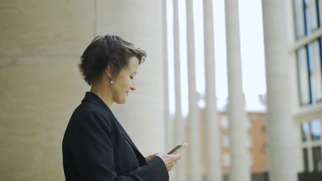 Elegant businesswoman standing outdoors next to office building, texr messaging on her cell phone and looking away with dreamy smile, full circle pan