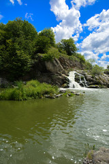 The waterfall on the river flows through and over the rocks covered with lichen and moss against a background of green vegetation and a blue sky.