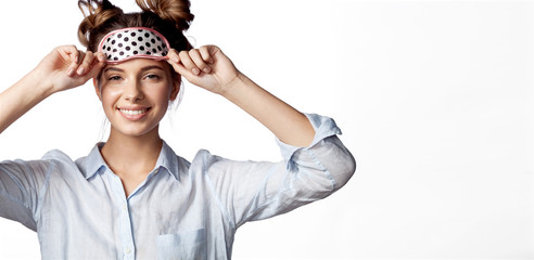 Portrait of cheerful young woman in white shirt posing at studio with eye pillow. Joyful female with amusing hairdo looking at camera with happiness and gladness