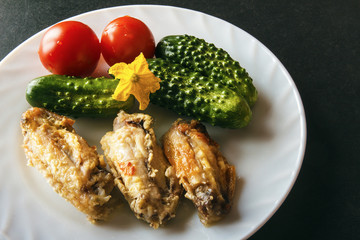 Baked chicken wings, fresh tomatoes and green cucumbers on a white plate on a black background. Dietary healthy food for dinner. Copy space