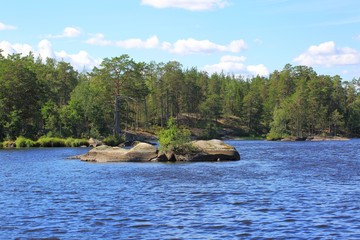 Nice view of rocky island in the middle of a lake. Blue lake water and green trees on blue sky with white clouds background. Sweden, Europe.