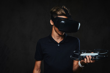 Young teenager dressed in a black t-shirt wears virtual reality glasses and holds a quadcopter. Isolated on a dark background.