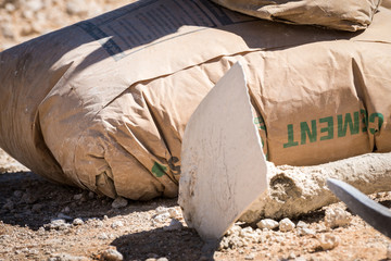 Unopened brown cement bag, a hoe and a pick axe, on the ground outside on a construction site