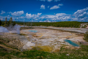 Pools of colorfully colored water dot the landscape of the Norris Geyser Basin in Yellowstone National Park with a mountains partial covered with snow behind