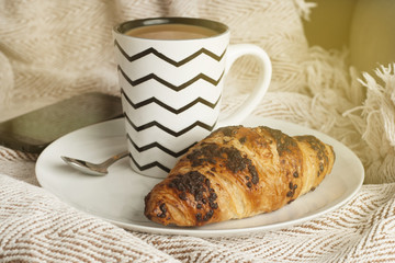 Cup of coffee with croissant on a white plate, with warm blanket. Breakfast, selective focus, toned.