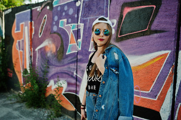 Stylish casual hipster girl in cap, sunglasses and jeans wear, listening music from headphones of mobile phone against large graffiti wall.