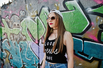 Stylish casual hipster girl in cap, sunglasses and jeans wear against large graffiti wall.