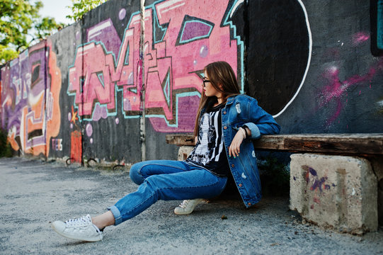 Stylish casual hipster girl in jeans wear and glasses against large graffiti wall.