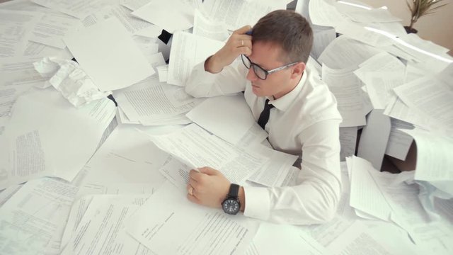 A young tired man in glasses sits in a pile of papers and holds his head. Due to the number of documents, an office worker can not cope with work and worries.