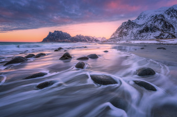 Beautiful sandy beach with stones in blurred water, colorful cloudy pink sky and snowy mountains at sunset. Utakleiv beach, Lofoten islands, Norway. Winter landscape with sea, waves, rocks at dusk - obrazy, fototapety, plakaty