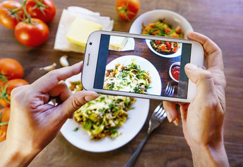 Hands with the smart phone pictures of meal. Young woman, cooking blogger is cooking at the home kitchen in sunny day and is making photo at smartphone. Instagram food blogger workshop concept.