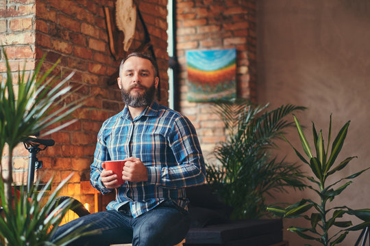 Handsome bearded hipster male in a blue fleece shirt and jeans holds a cup of morning coffee while sitting on a wooden stool at a studio with a loft interior.
