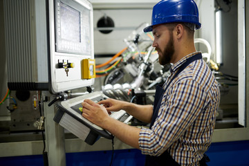 Side view portrait of machine operator wearing hardhat sitting at control panel pressing buttons,...