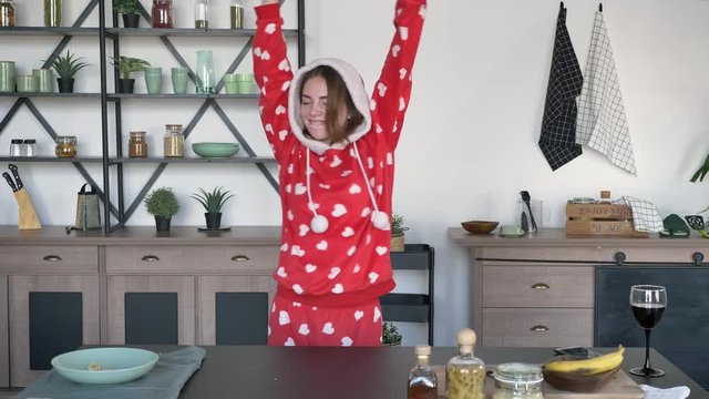 Happy ginger woman wearing hood of funny pajamas with hearts and dancing in modern kitchen behind cooking table