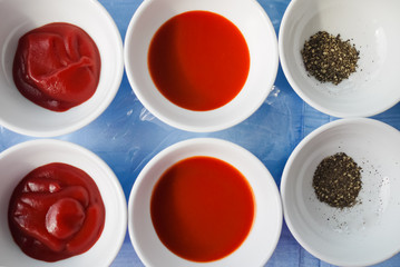 Assortment of basic food flavoring and seasoning ingredients ready for cooking. Ketchup, Red Chili Sauce and Ground Black Pepper in separate bowls on blue table. Close up. Natural light.
