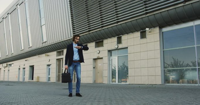 Young handsome man in business style standing outdoor and talking on the phone, then looking at the watch and started to tun and rush as being late.