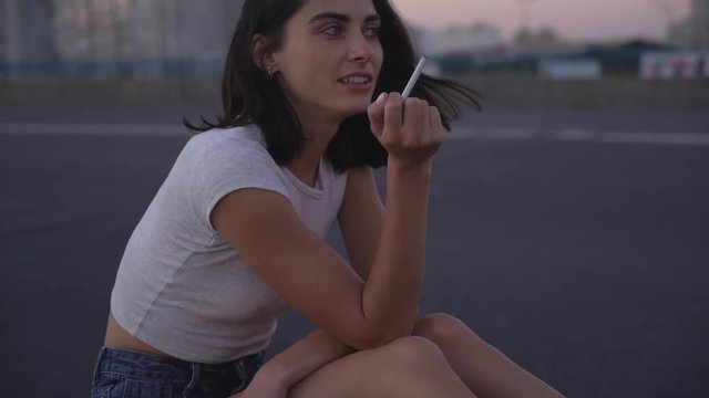 cute sexy brunette smokes a cigarette. A woman sits on the side of the highway and smokes thoughtfully. Warm summer evening 4K