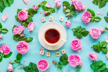 Fototapeta na wymiar Top view creative layout with Tea time lettering with wooden blocks, cup of hot tea and fresh pink tea rose flowers, buds, petals, leaves on the blue background. Flat lay. Selective focus. Copy space.