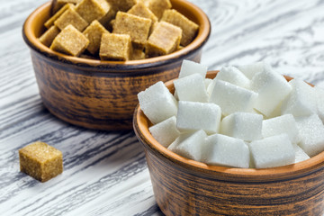 White refined sugar and brown unrefined sugar cubes in ceramic bowls on a light wooden table.