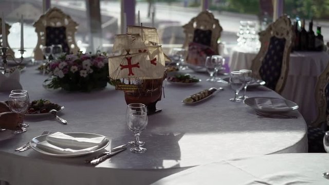 Plate, cloth napkin, knife and fork on white cloth on table in luxury restaurant decorated with model of old wooden ship, candles and flowers composition