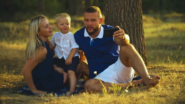 Happy family is sitting on the grass and doing selfie with a baby at sunset in the park. Father and mother take pictures of themselves with the baby on the phone