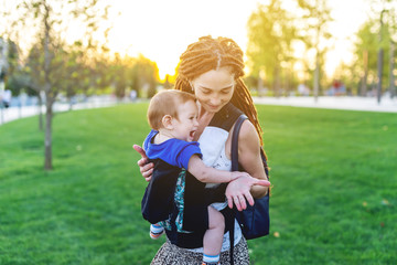 Young happy mom with baby son in ergo backpack travel together. Sunny summer day. Concept of modern mother