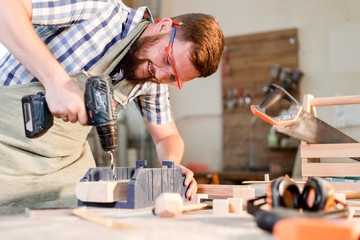 Bearded carpenter with safety glasses drills a wooden bar