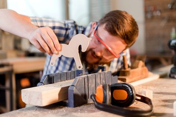 Bearded carpenter in goggles playing with a wooden figurine in a carpentry workshop