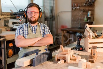 Bearded carpenter in goggles standing in the carpentry workshop with arms crossed