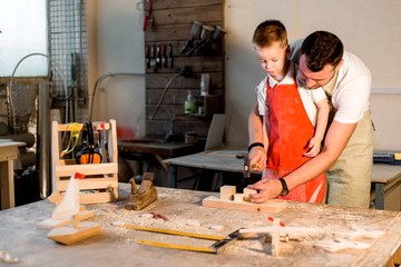 Dad teaches his son carpentry craft in the carpenter's shop hammering nails into a wooden bar