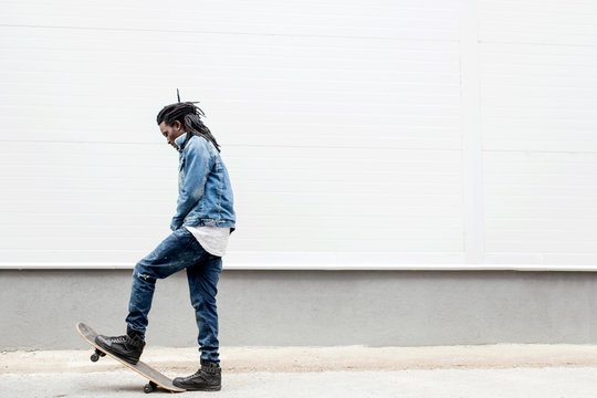 African man with dreadlocks in jeans jacket rides a skateboard on the background of the building