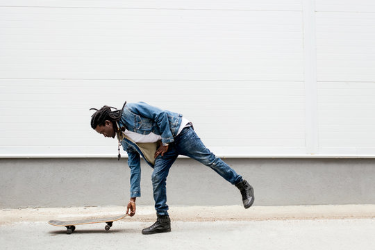 African man with dreadlocks in jeans jacket rides a skateboard on the background of white walls - Powered by Adobe