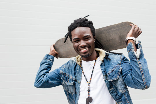 African man with dreadlocks in a denim jacket holding a skateboard on his shoulders closeup