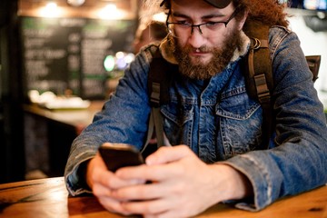 Hairy man traveler in a denim jacket with a backpack sitting at a table in a cafe with a phone in his hands
