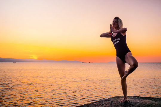 A beautiful girl in a black swimsuit with the inscription "Palm beach Florida" does yoga on the pier by the sea, standing asana Vrikshasana at dawn. Healthy lifestyle.