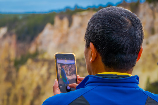 Close up of selective focus of man using a cellphone to take a picture of a friend posing in front o the upper Yellowstone Falls in Yellowstone National Park, Wyoming, United States