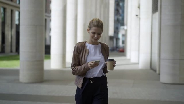 Dolly shot of smiling young businesswoman with takeaway coffee cup in her hand walking down street and texting on her smartphone