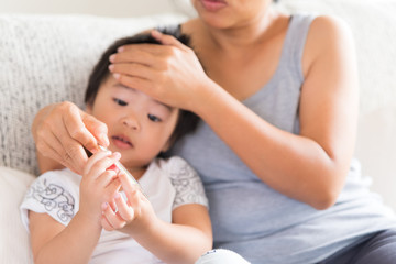 Close up of a mother checking the temperature of her ill baby with a thermometer on a couch in the living room at home. Medicine and health care concept.