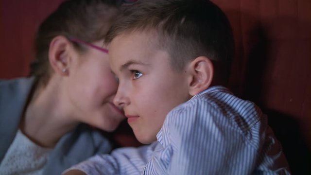 Girl whispering to boy watching movie in cinema. Children exchange impressions about film. Close up of boy watching cinema movie in slow motion. Child movie entertainment