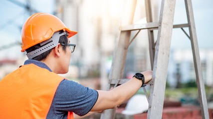 Young Asian maintenance worker man with orange safety helmet and vest carrying aluminium step...