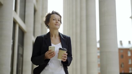 Medium dolly shot of elegant middle aged businesswoman with short hair walking down street with cell phone and takeaway coffee cup in her hands and looking around thoughtfully - Powered by Adobe