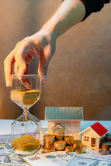 Real estate or property investment growing business. Home mortgage loan rate. Saving money for retirement concept. Male hand holding hourglass with house model and coin stack on banknotes on the table