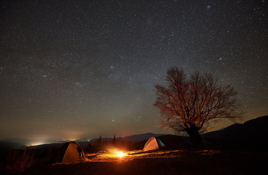 Fantastic night camping site view. Bright bonfire burning between two tourist tents under beautiful dark starry sky on big tree and distant mountain range background. Tourism, outdoor activity concept