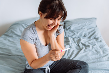 young happy woman sitting on bed looking on positive pregnancy test