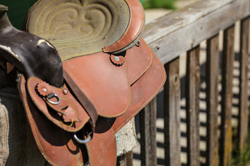 Old worn leather horse saddle on wooden fence, lit by sun.