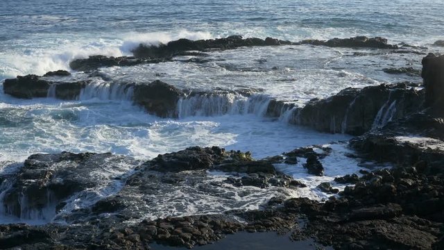 Slow motion (59.94 fps rendered at 29.97), zooming out view of a wave crashing against, and cascading down, lava rock, near Spouting Horn blowhole in the Koloa District of southern Kauai, Hawaii.