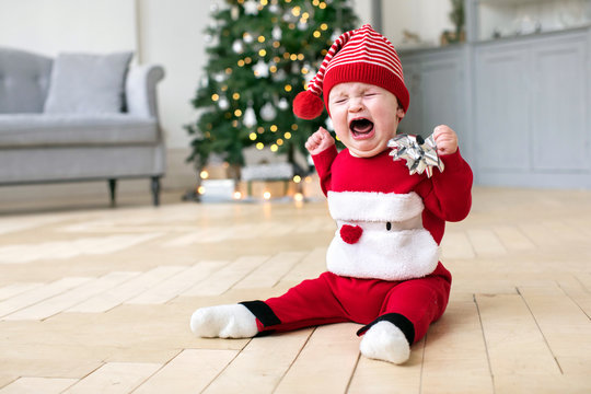 Cute baby in red Christmas costume sitting on floor of stylish room and crying